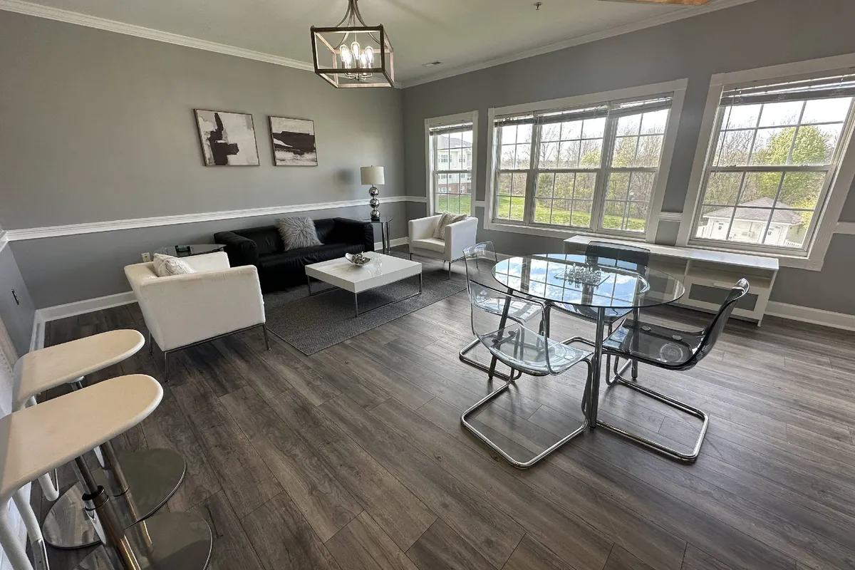 A spacious and sunlit open living area featuring a cozy sofa, a dining table set for gatherings, large windows that invite in natural light, and tastefully chosen artwork that adds a touch of personality to the space. The room exudes a contemporary and welcoming atmosphere, perfect for both relaxing and socializing.