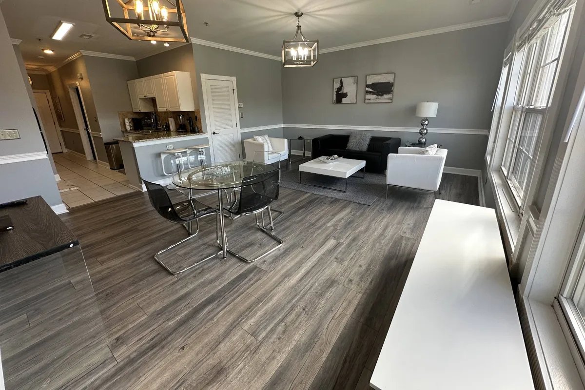 A sunlit dining and lounge space with a sleek black sofa, a coffee table, a dining table with chairs, and large windows that allow natural light to highlight the inviting atmosphere. The setting is ideal for intimate dinners, socializing, or simply relaxing after a day exploring Charlotte.
