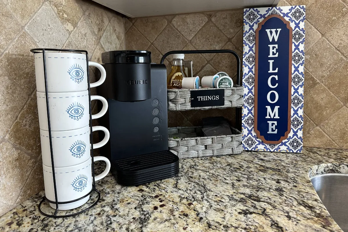 A close-up of a kitchen coffee station, featuring a modern coffee maker, a stack of cups, a friendly welcome sign, and a basket of amenities. The cozy vignette creates a sense of daily ritual and hospitality, emphasizing home-like comfort.