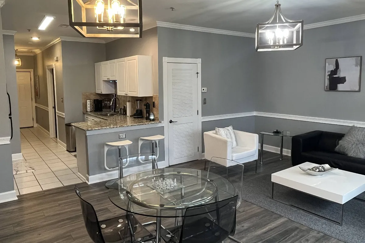 A contemporary interior scene showing a sleek glass dining table, black sofa, inviting white armchair, and a view of the adjacent kitchen. The space feels airy and cohesive, designed for both relaxation and hosting—ideal for entertaining guests or enjoying quiet moments. The subtle luxury touches are evident in the finishes and furnishings.