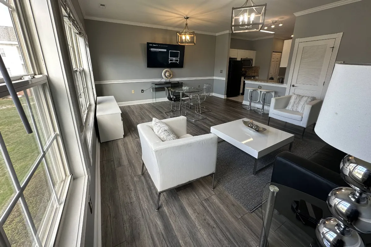 A modern yet cozy living and dining space highlighted by sleek white armchairs, a stylish glass dining table, an eye-catching light fixture, and a TV mounted on the wall. The area is set up for both social occasions and peaceful mornings, reflecting a blend of functionality and style.