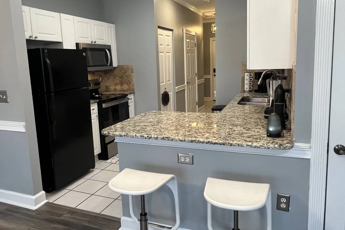 A bright, modern kitchen featuring granite countertops, sleek black appliances, white cabinetry, and a row of bar stools at the breakfast bar. The space is uncluttered and inviting, ideal for both meal prep and casual dining.
