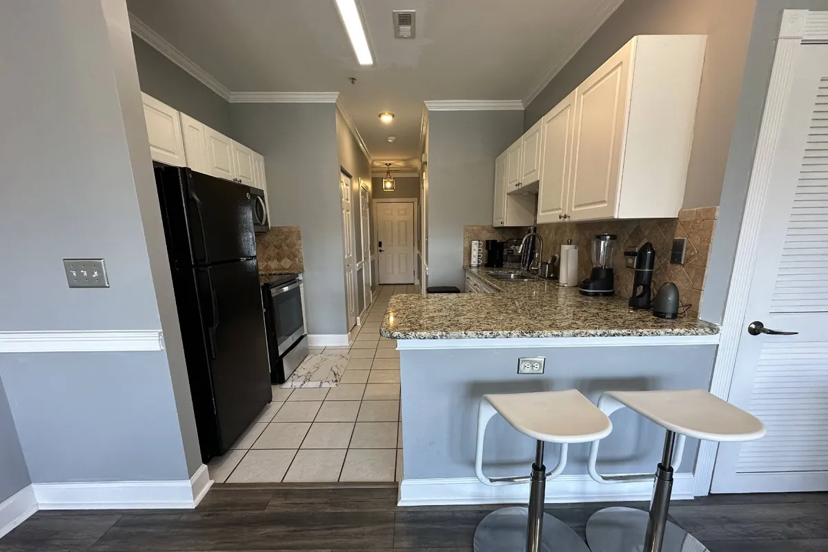 A welcoming kitchen scene with granite countertops, black appliances, white cabinetry, and comfortable bar stools. The setup is ideal for both meal preparation and socializing, suggesting an environment where cooking and connection blend seamlessly.
