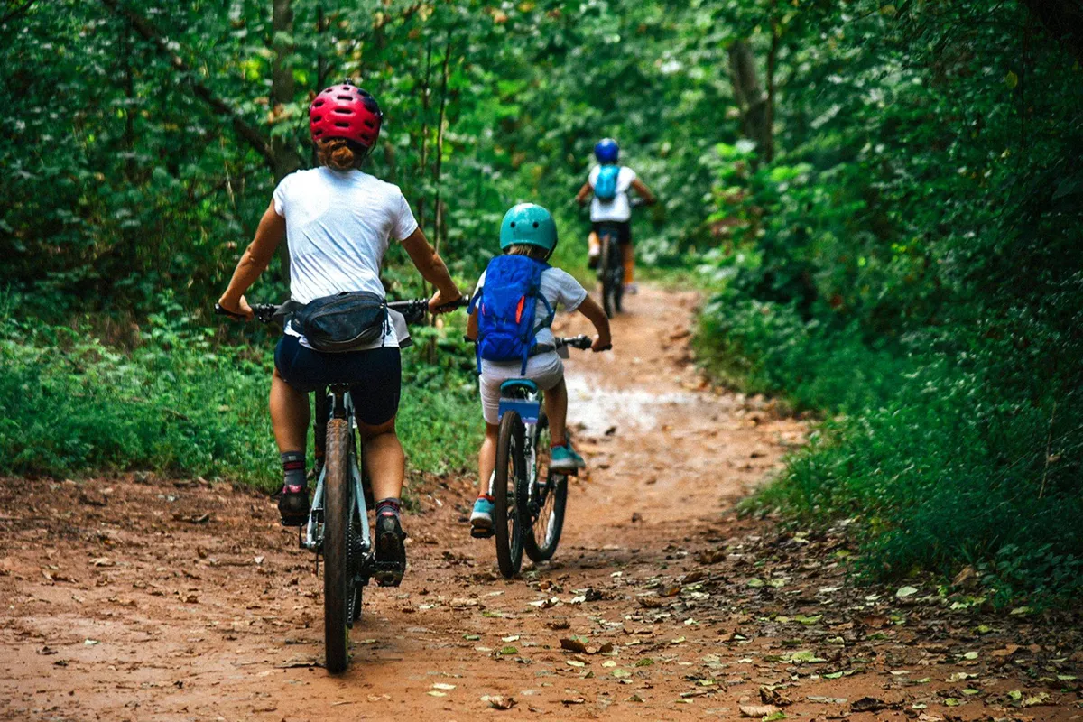 An exterior scene of a family biking together along a tree-lined dirt path, surrounded by lush greenery. The joyful and adventurous mood suggests active exploration, family outings, and access to nature trails or local parks.
