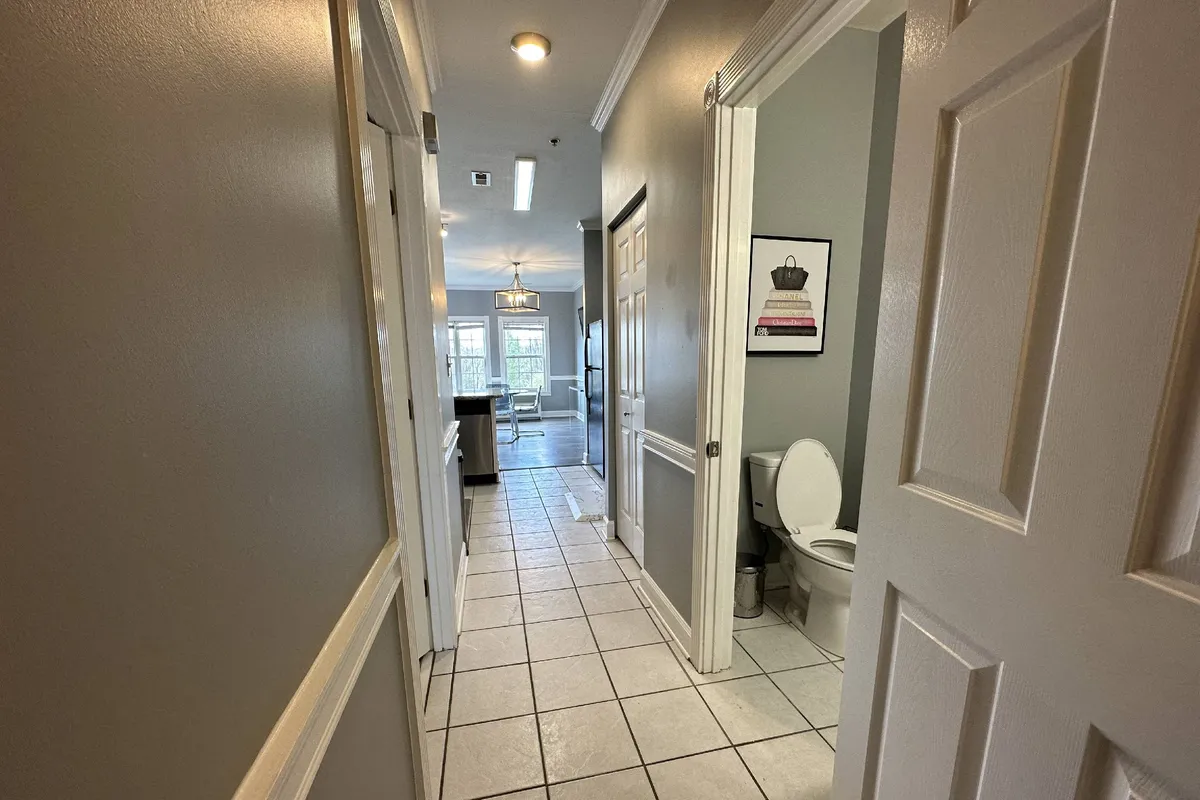 A view down a bright hallway leading to the kitchen, showing framed artwork, a bathroom door, and a glimpse of the living space. The scene exudes warmth and functionality, supporting both social and private moments within the condo.