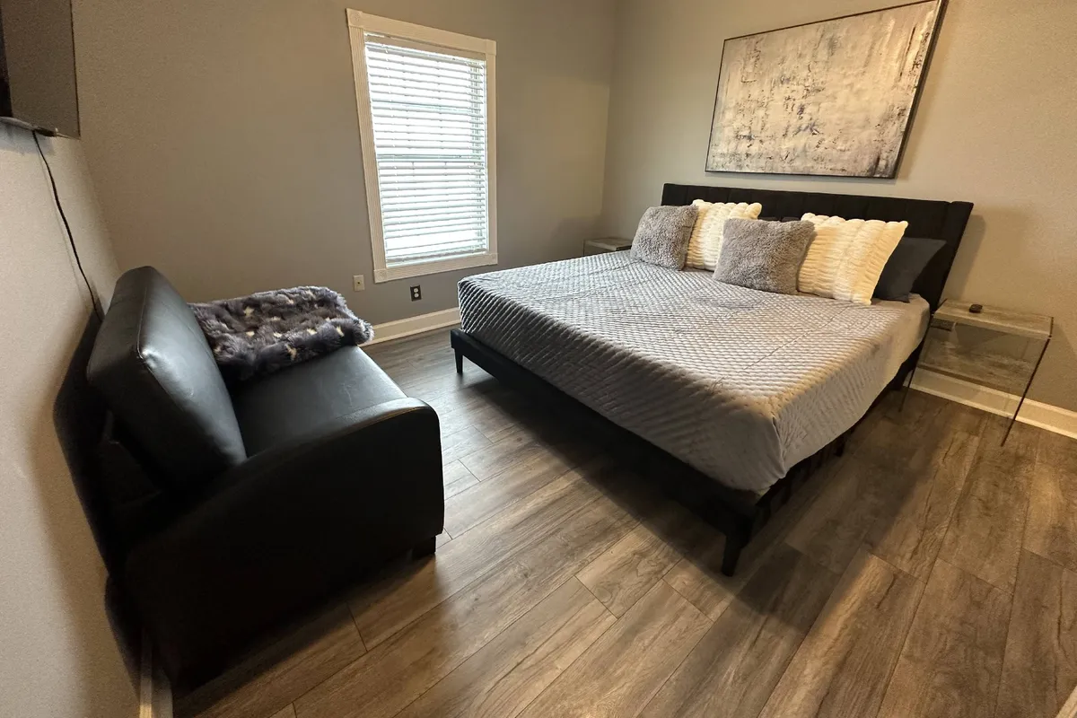 This cozy interior scene features a comfortable bed, a sofa for lounging, tasteful artwork, and a window letting in gentle daylight. The arrangement invites relaxation, quiet reading, or enjoying intimate moments, underscoring the room's adaptability for various needs.