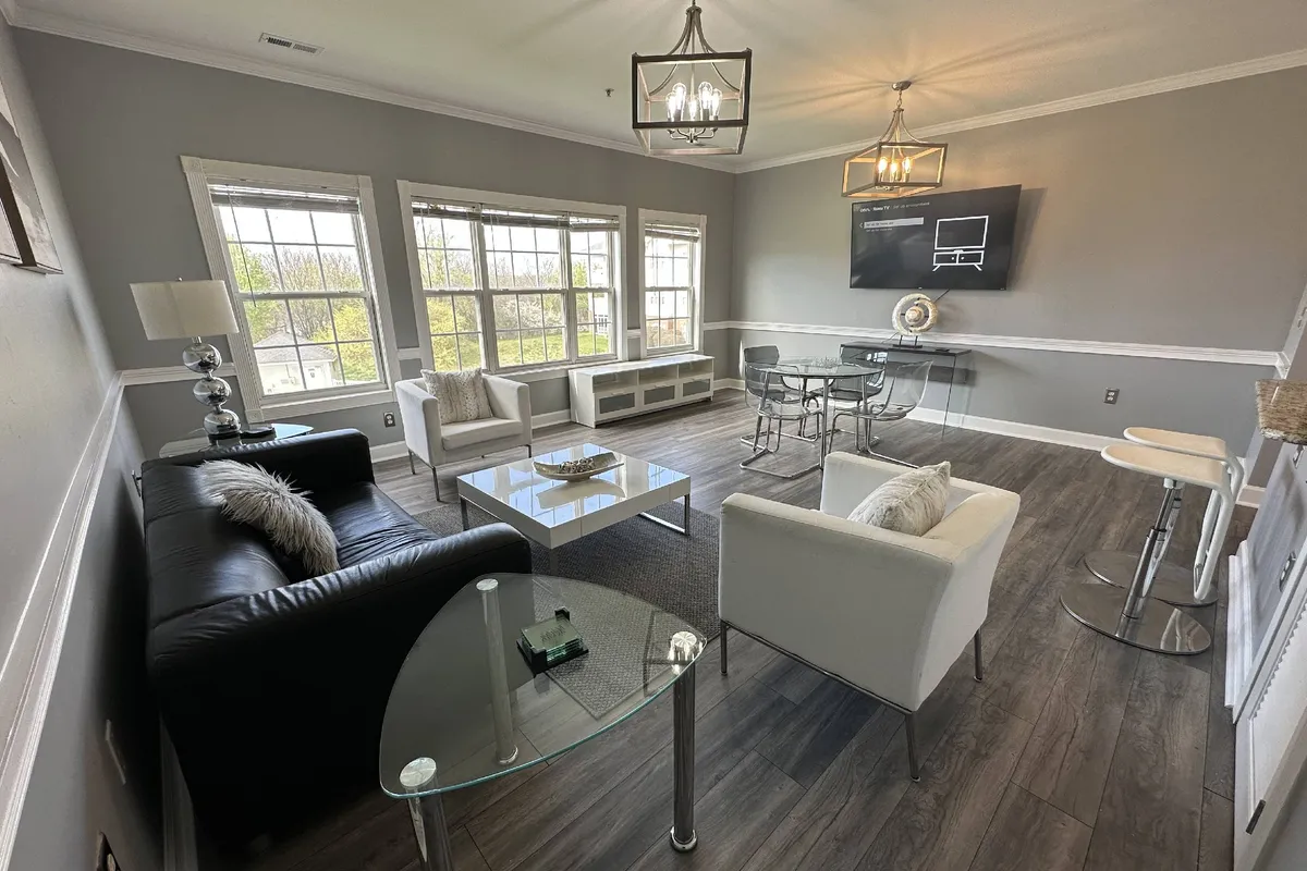The image shows a beautifully remodeled condo interior with large windows that flood the space with natural light, modern furniture arranged for socializing or relaxation, stylish light fixtures, and a sleek glass dining table perfect for dining, working, or gathering with friends.